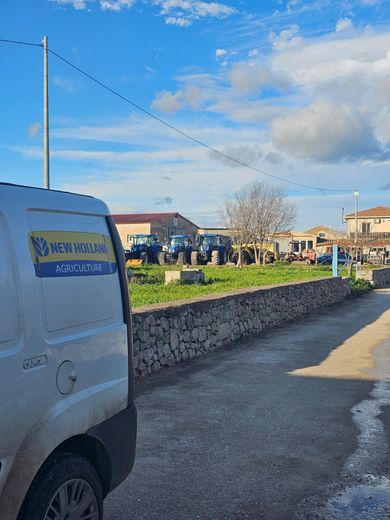 Scena di strada con un furgone bianco, un muro di pietra e persone riunite vicino alle case sotto un cielo azzurro.