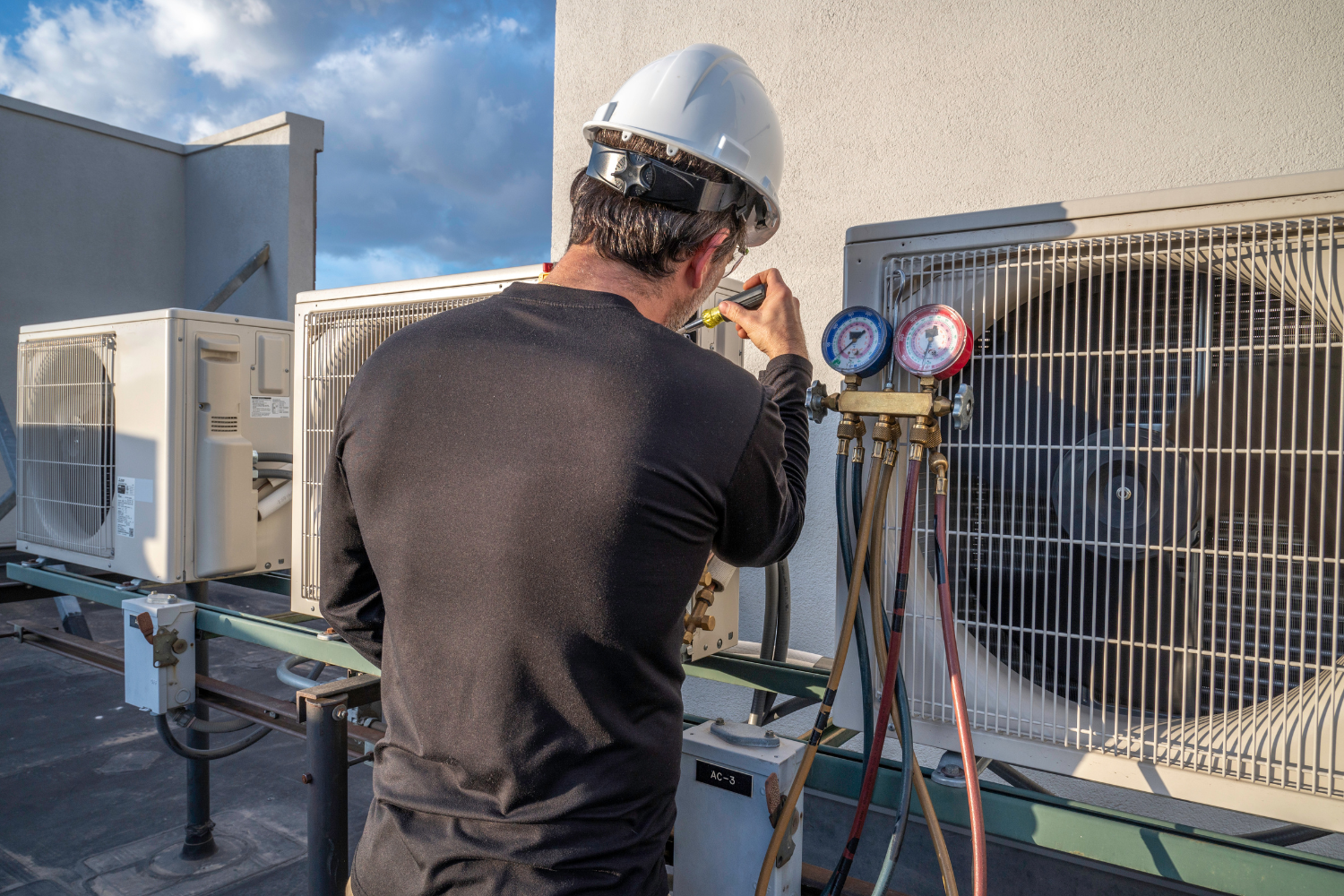 HVAC technician in hard hat repairing an air conditioning unit on a rooftop, using gauges and hoses.