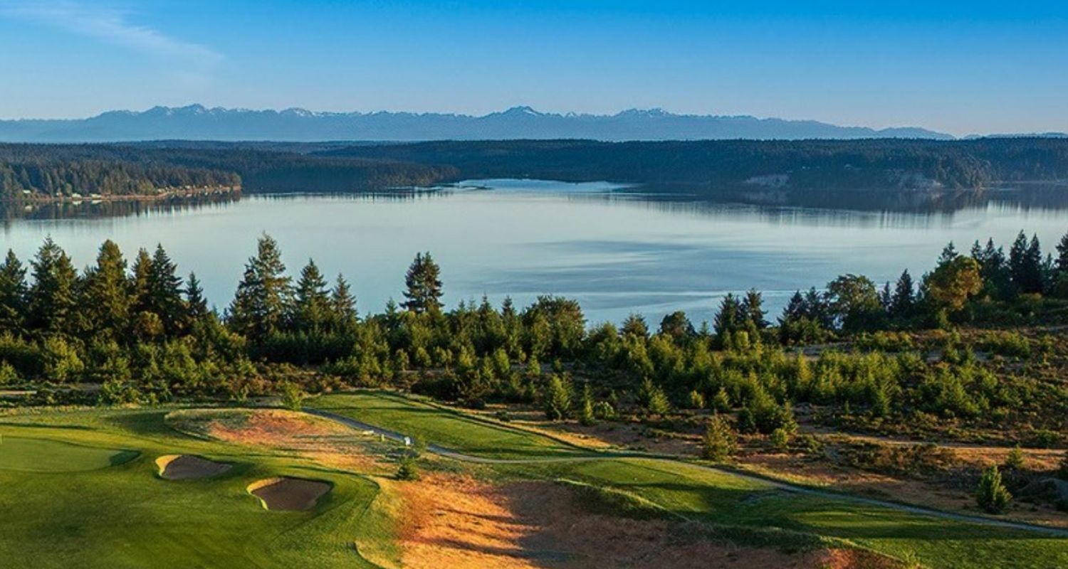Lush golf course with green fairways, sand traps, and a body of water with distant mountains under a blue sky.