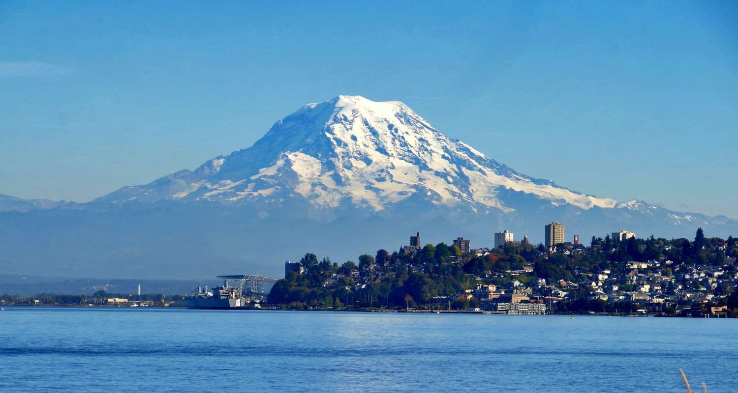 Snow-capped mountain, possibly Mount Rainier, over a body of water with a shoreline of buildings under a clear blue sky.