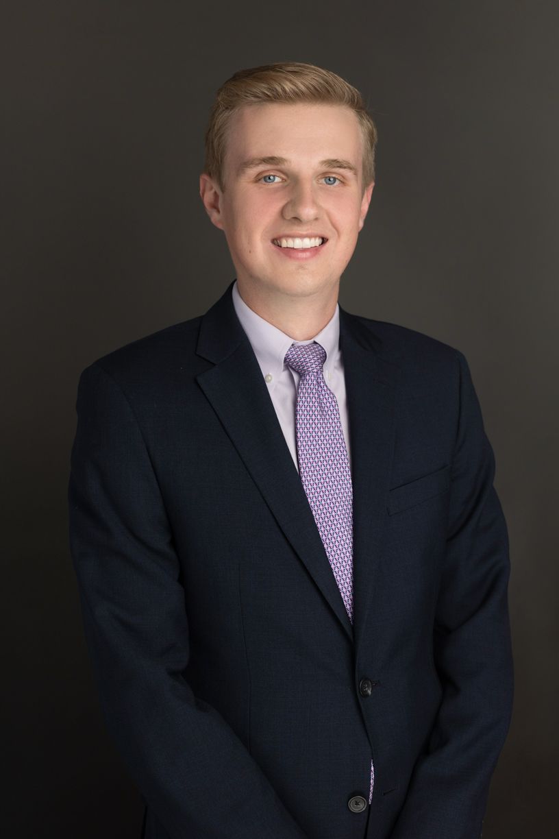 Young man with blond hair in a navy suit, light purple shirt and patterned tie, smiling against a dark gray background.