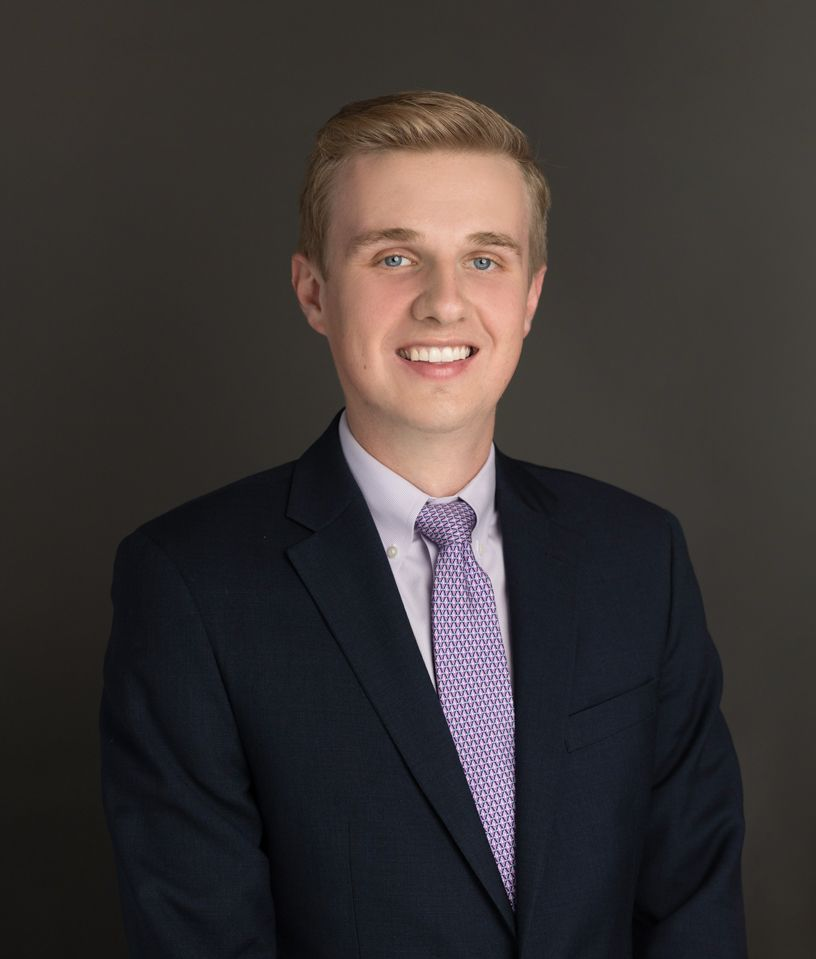 Blond man in a navy suit and purple tie smiles against a gray backdrop.