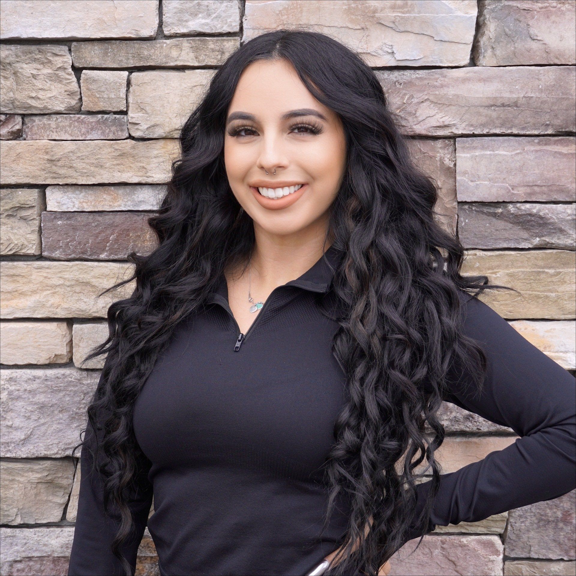 A woman with long black hair is smiling in front of a stone wall.