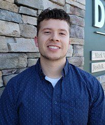 A young man in a blue shirt is smiling in front of a stone wall.