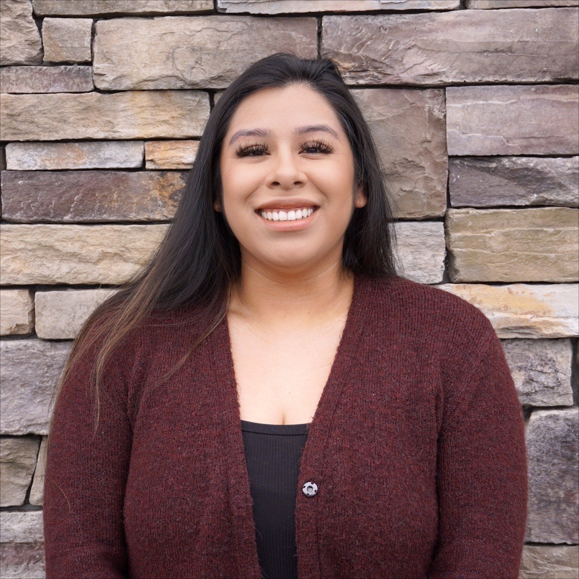 A woman in a maroon sweater is smiling in front of a stone wall.
