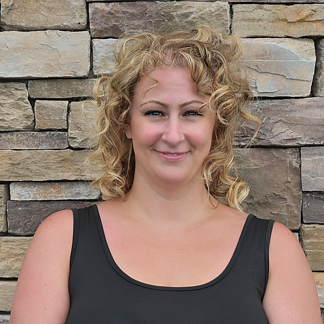 A woman with curly hair is smiling in front of a stone wall.