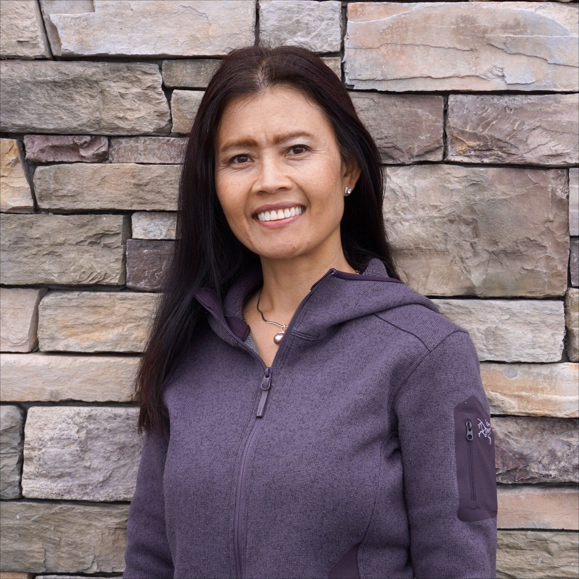 A woman in a purple jacket is smiling in front of a stone wall.