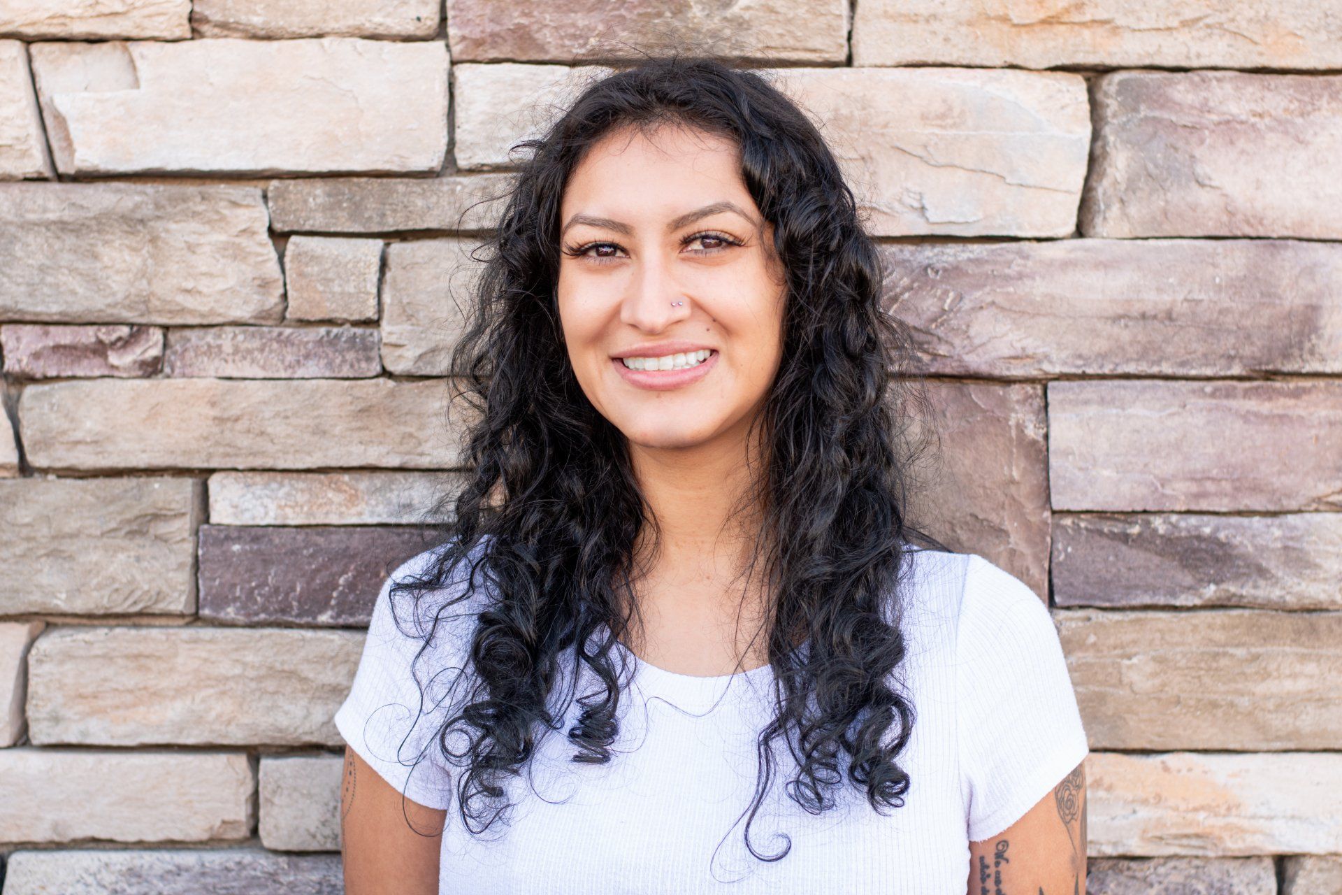A woman with curly hair is smiling in front of a brick wall.