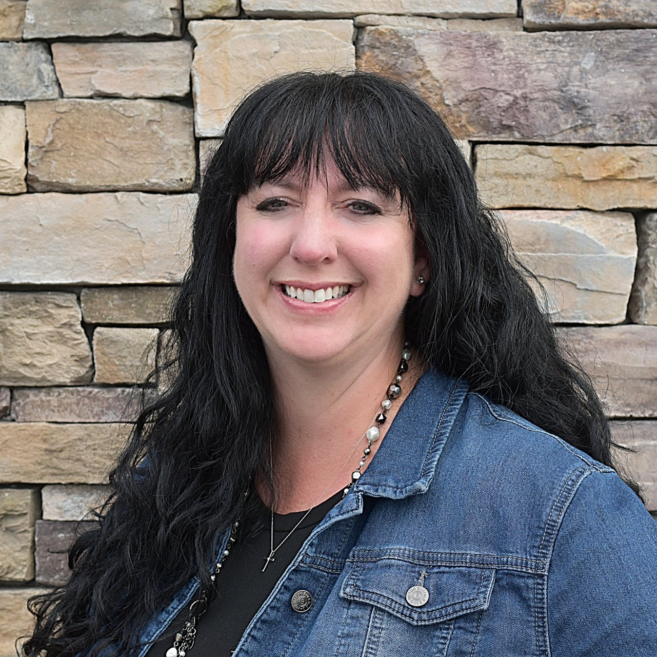 A woman in a denim jacket is smiling in front of a stone wall.