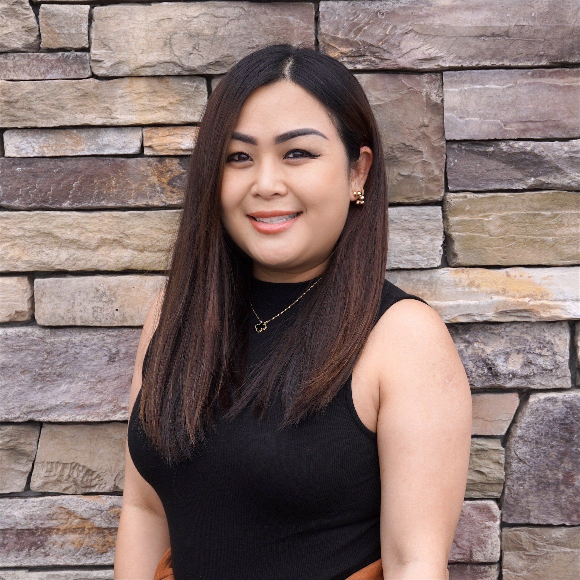 A woman in a black tank top is smiling in front of a stone wall