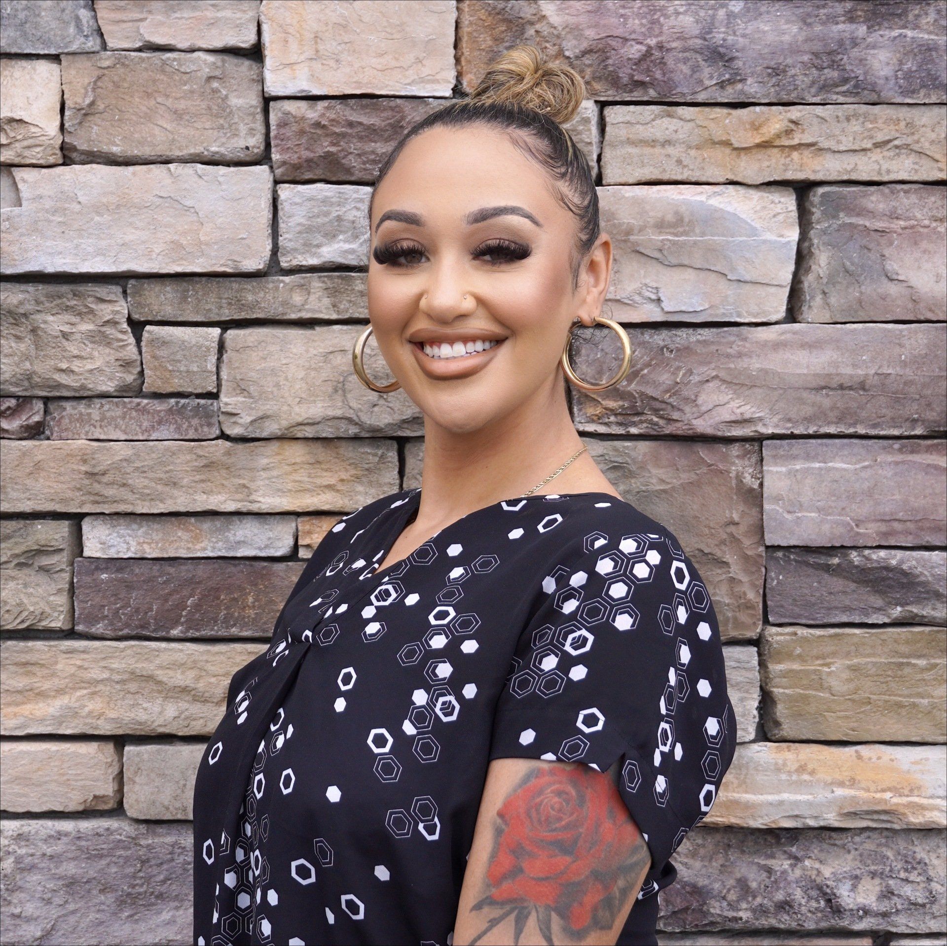 A woman with a tattoo on her arm is smiling in front of a stone wall.