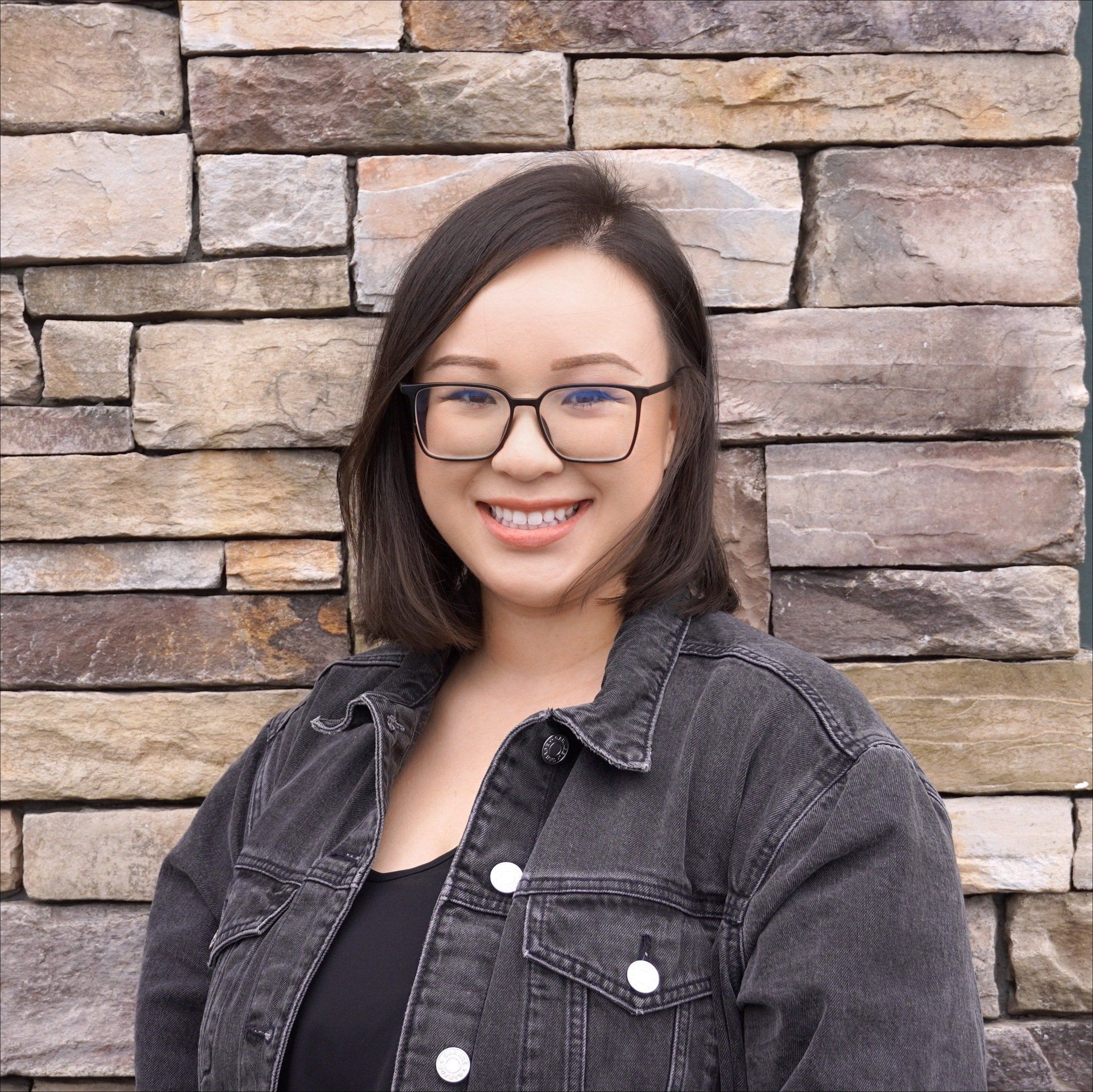 A woman wearing glasses and a denim jacket is smiling in front of a stone wall.