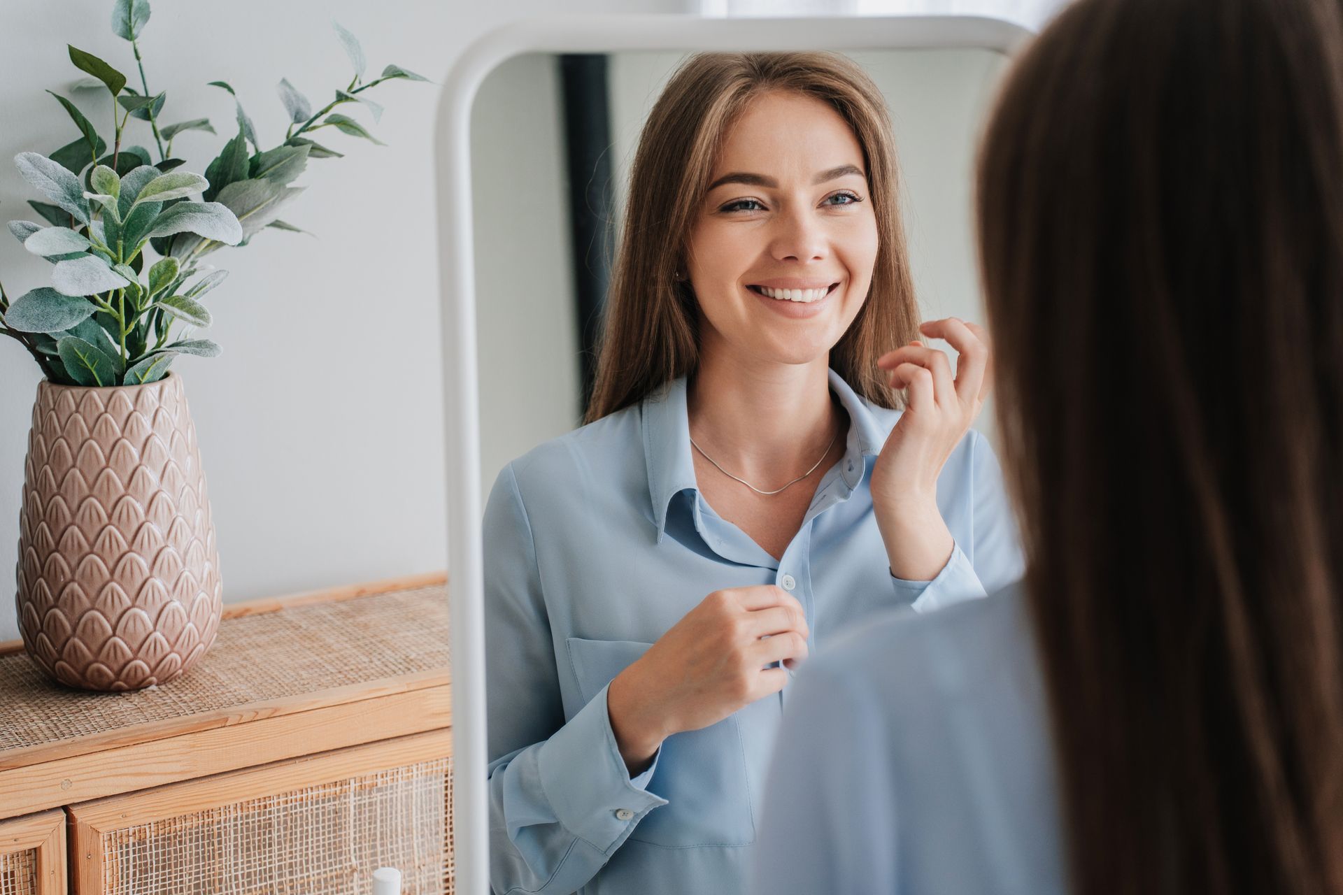 Woman smiling and adjusting her light blue blazer in a mirror beside a vase of flowers