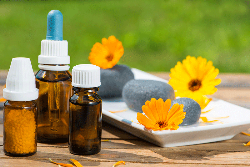 Ein Holztisch mit Flaschen homöopathischer Medikamente, Blumen und Steinen darauf.