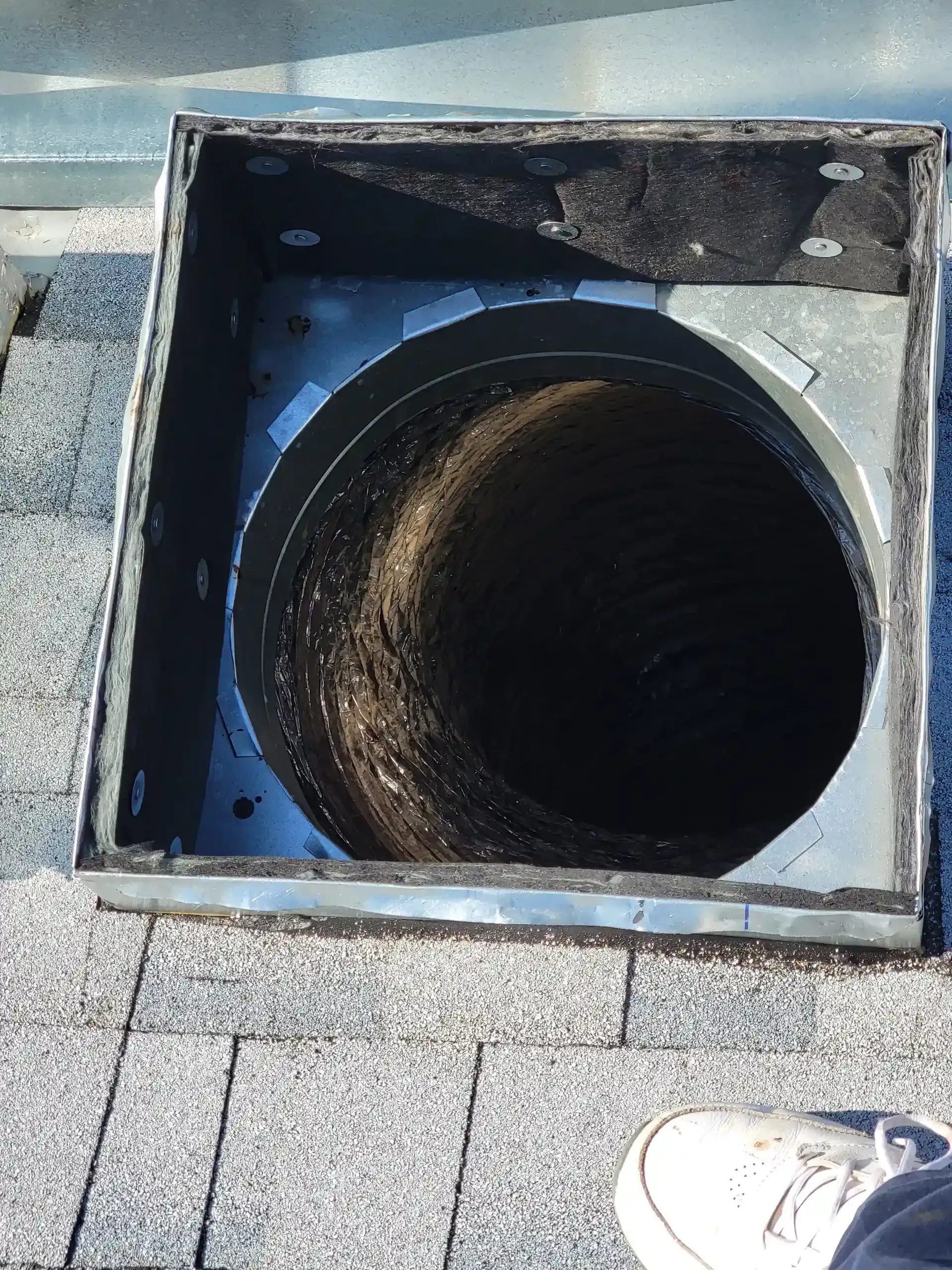A metal chimney box housing a circular flue opening on a shingled roof, viewed from above.