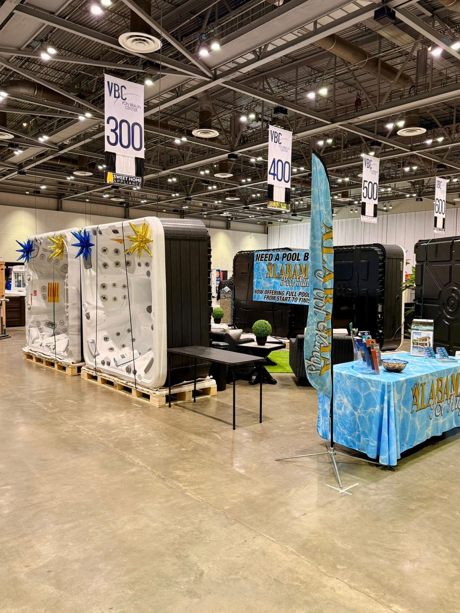 Trade show booths in a convention hall, with white and blue displays, hanging price signs, and polished concrete floor.