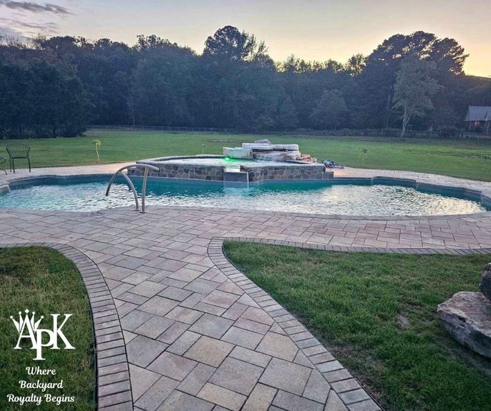 Backyard pool with brick patio, grassy lawn, and trees at dusk