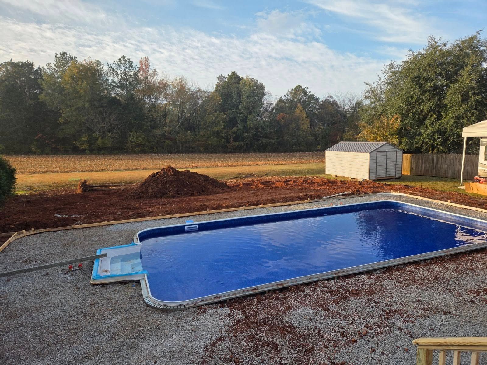 Backyard with an above-ground pool, gravel patio, and trees beyond a dirt yard