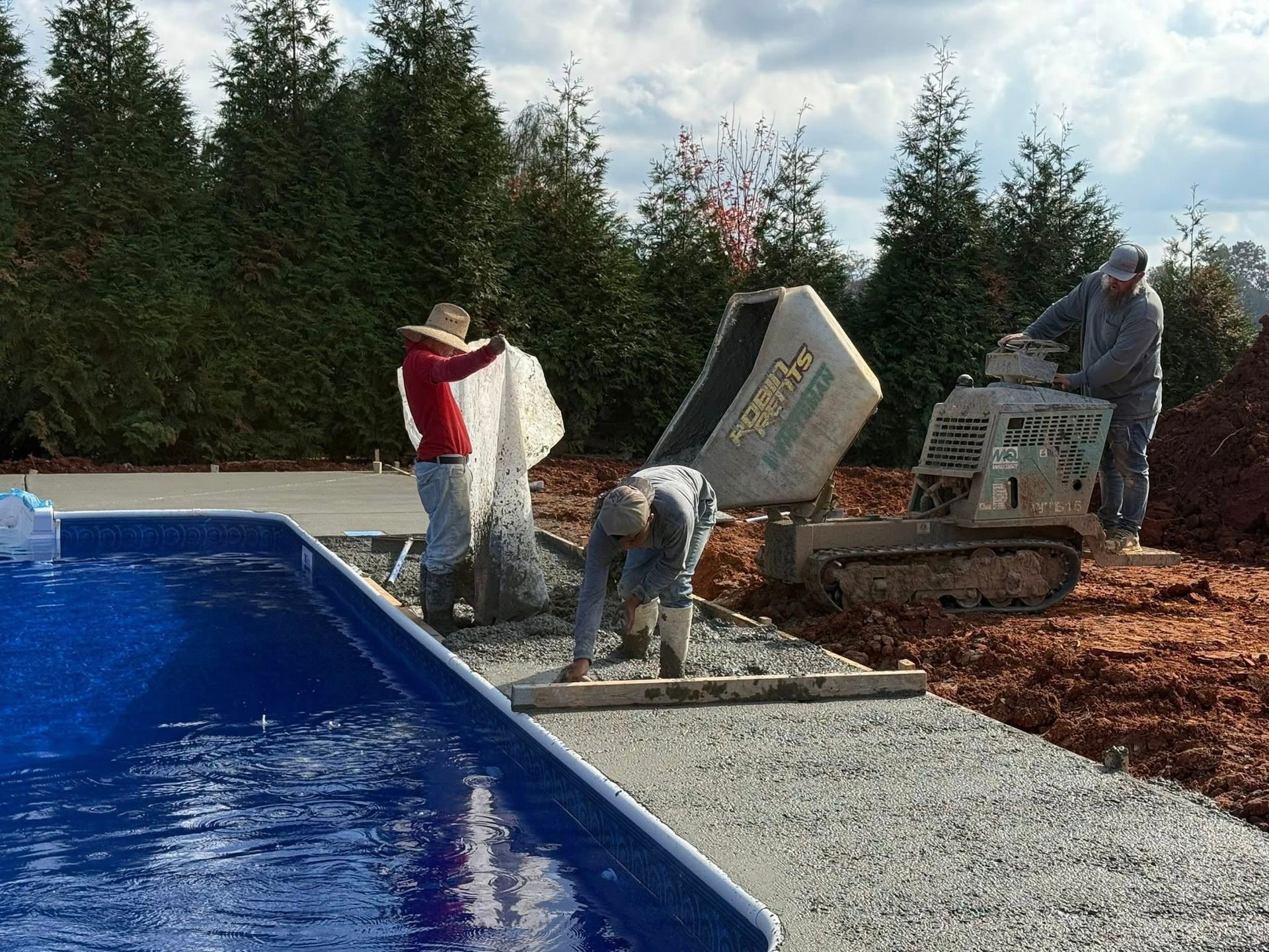 Workers installing a pool edge beside a blue swimming pool, with gravel and trees in the background