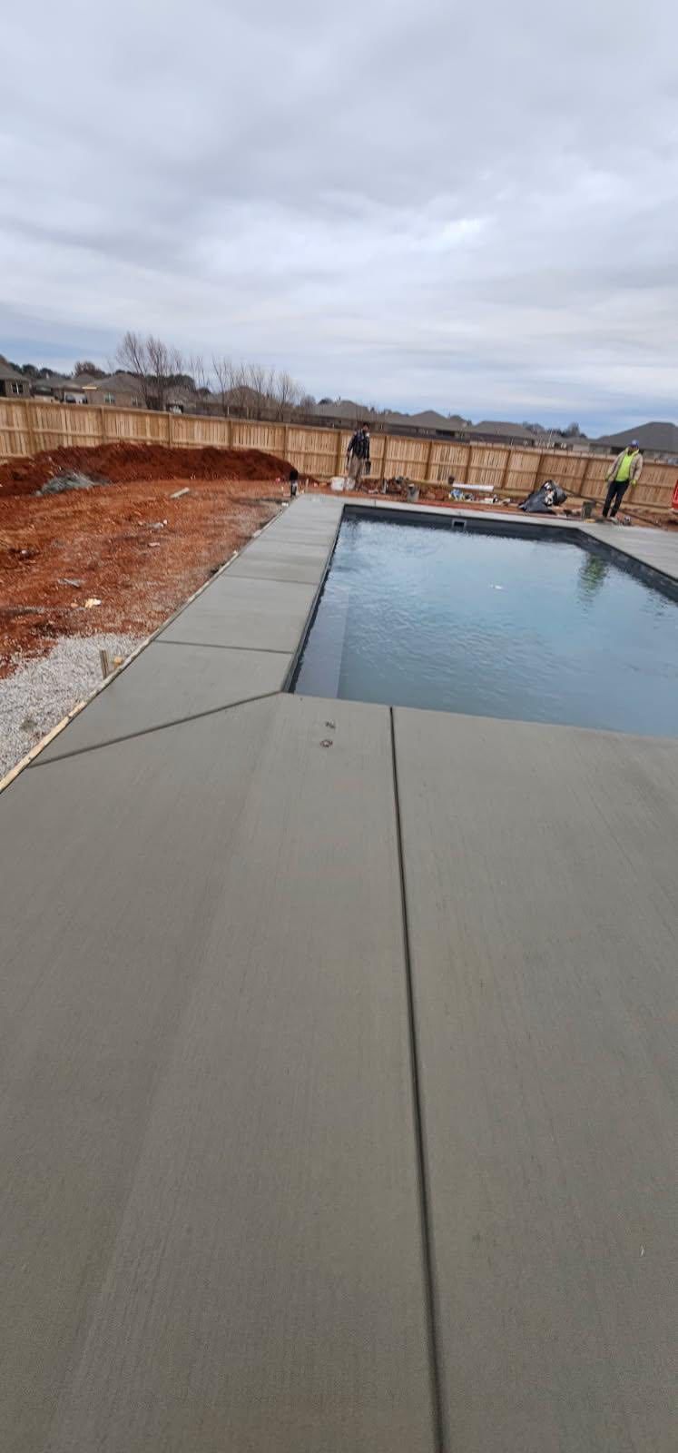 Partially completed backyard pool with concrete deck and cloudy sky over a fenced lot