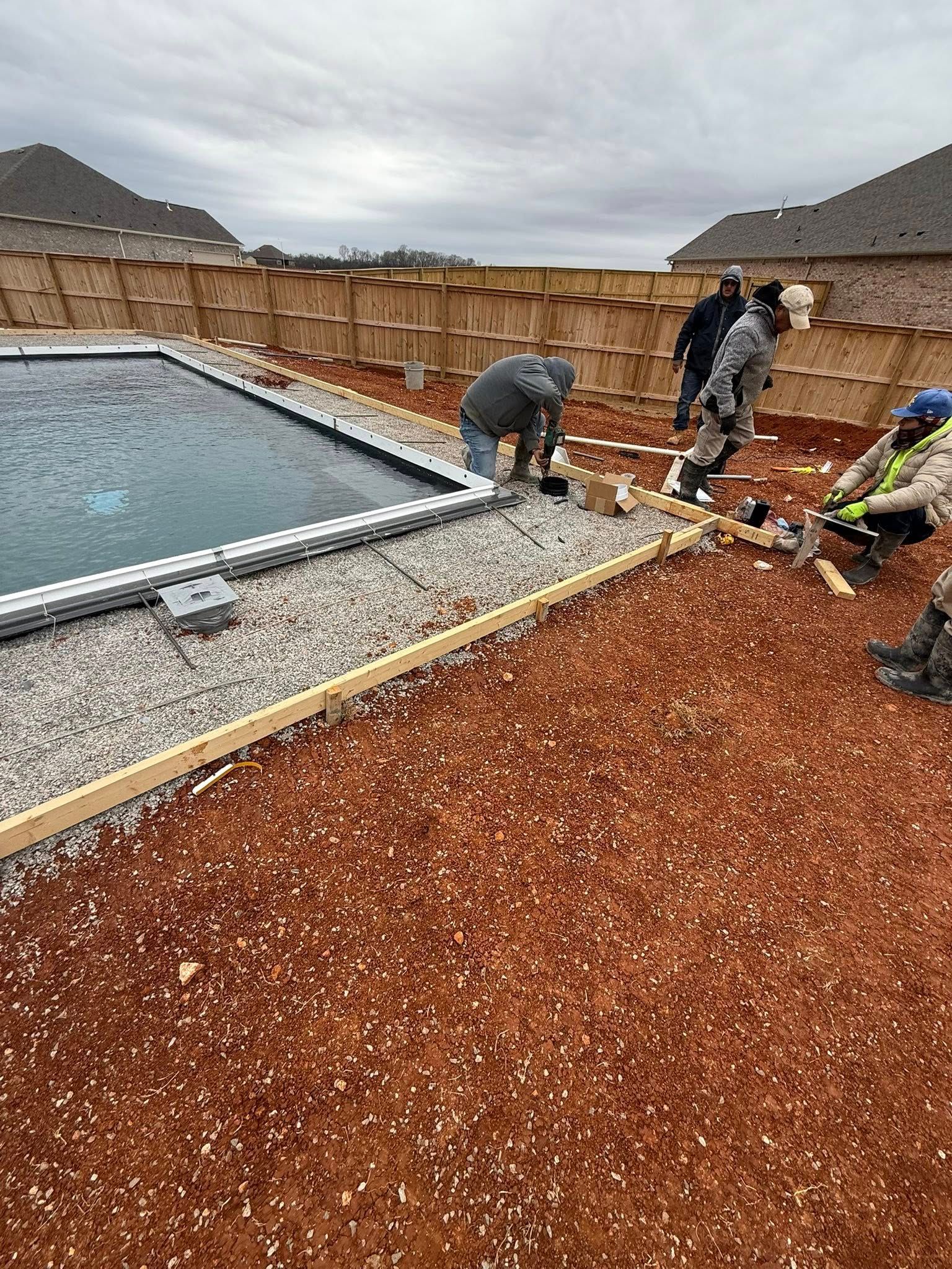 Workers installing pavers beside a backyard pool and gravel patio, with a wooden fence in the background