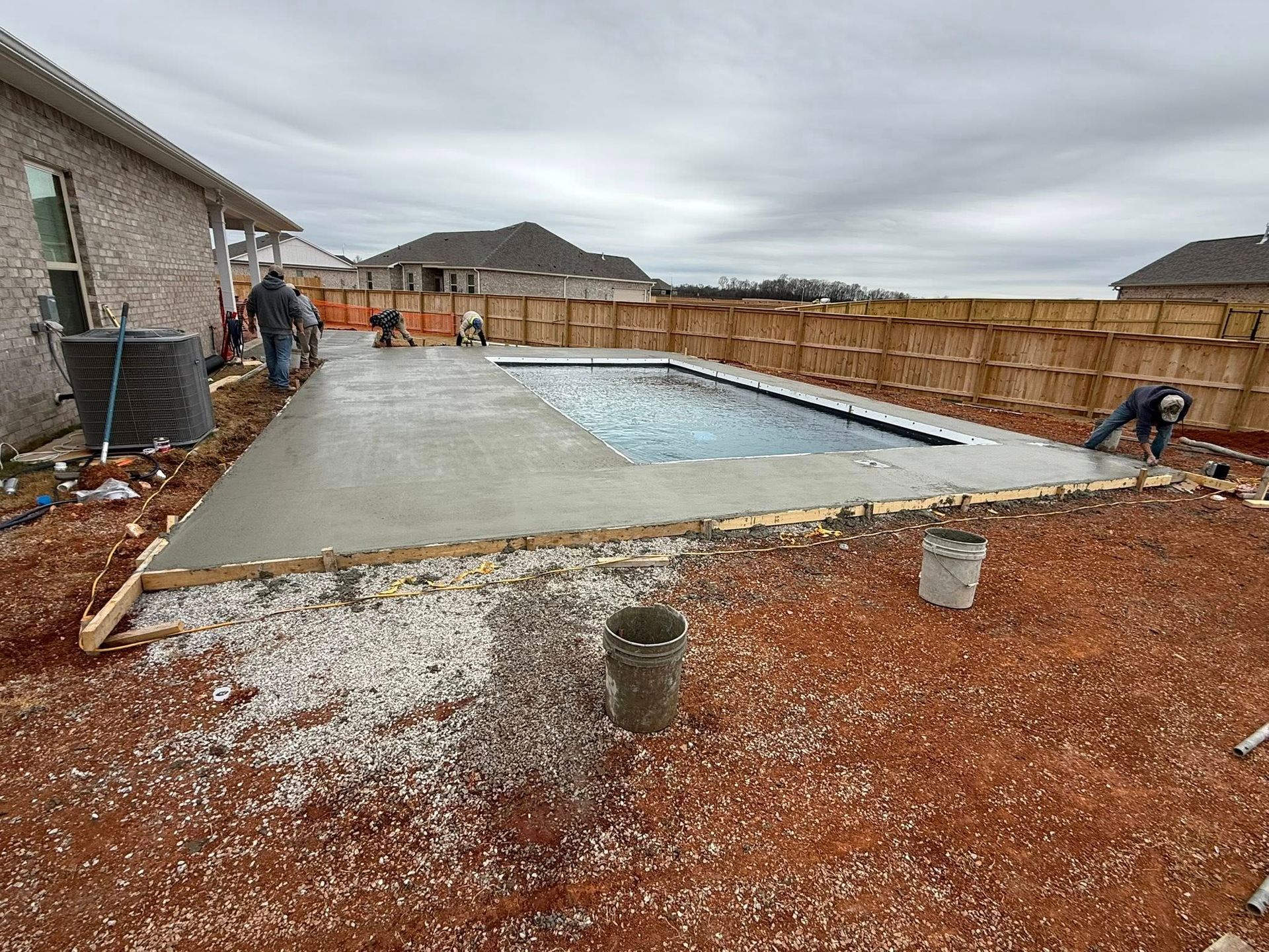 Backyard pool under construction beside a house, with workers, concrete deck, and wooden fencing.