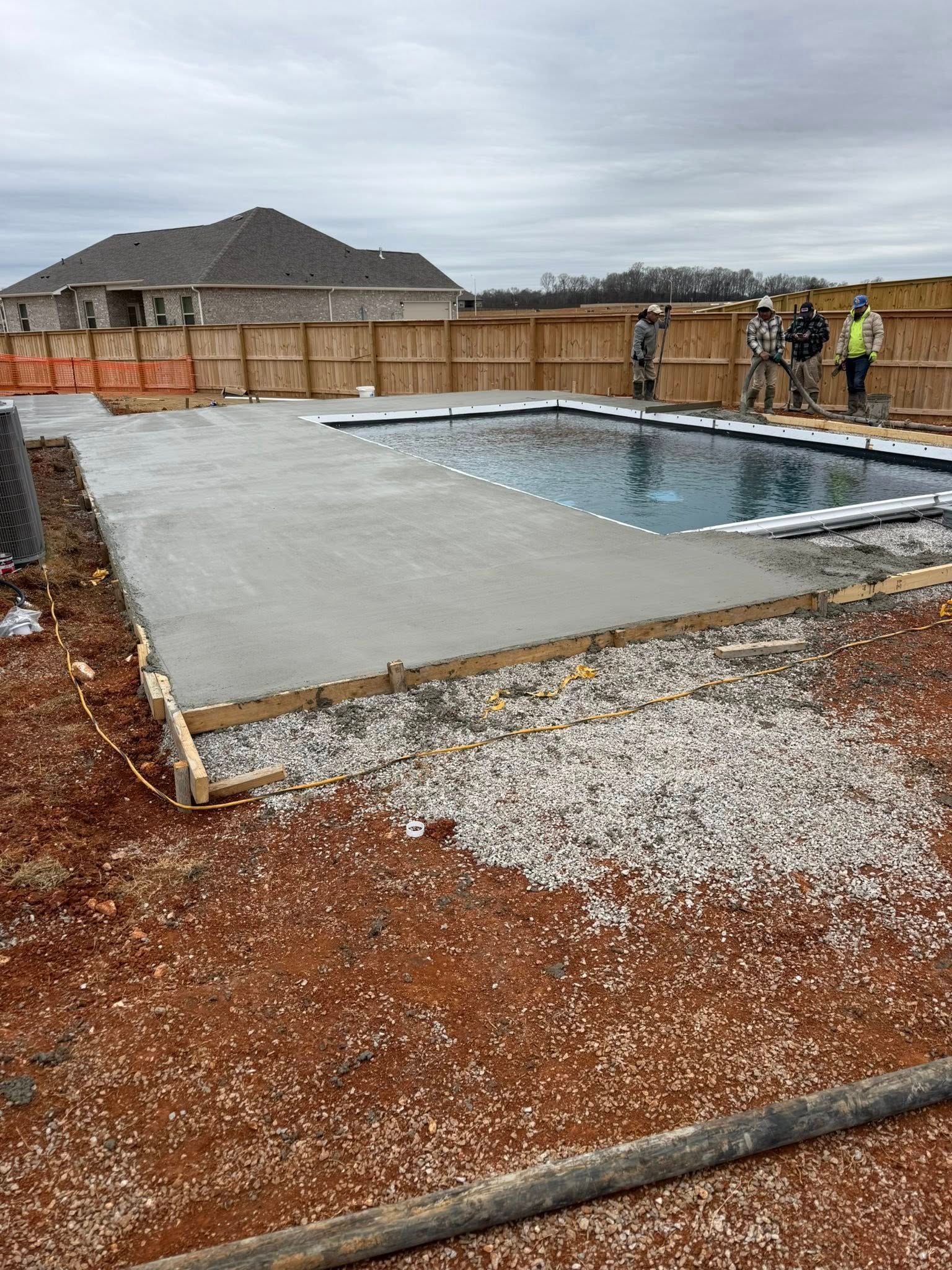 Partially poured concrete pool deck beside an in-ground pool under construction, with workers in the background