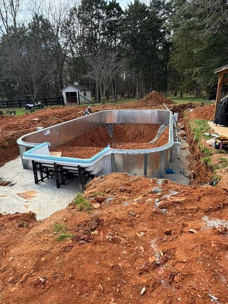Partially built in-ground pool surrounded by dirt and construction debris in a wooded yard