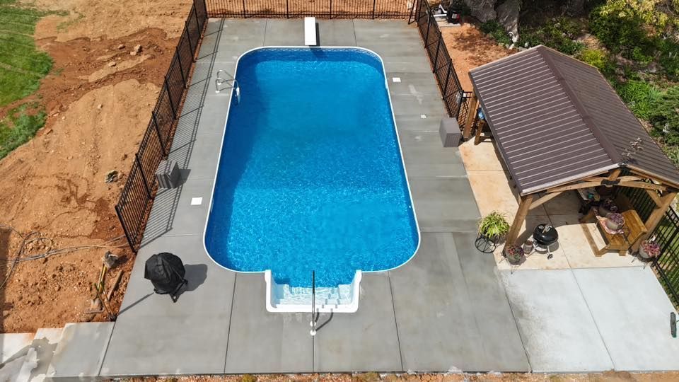 Aerial view of a rectangular blue swimming pool beside a patio and gazebo on a concrete deck.