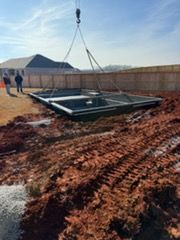 Crane lifting a black steel frame into a muddy construction site under a blue sky