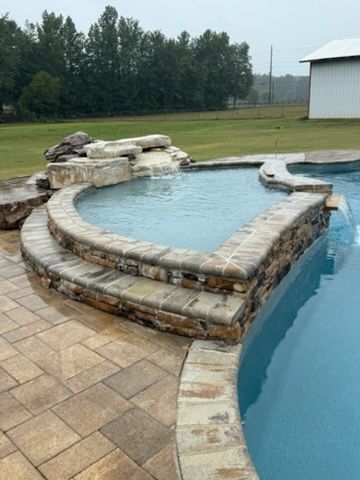 Stone pool with rock waterfall edge beside a larger pool on a patio, backed by grass and trees