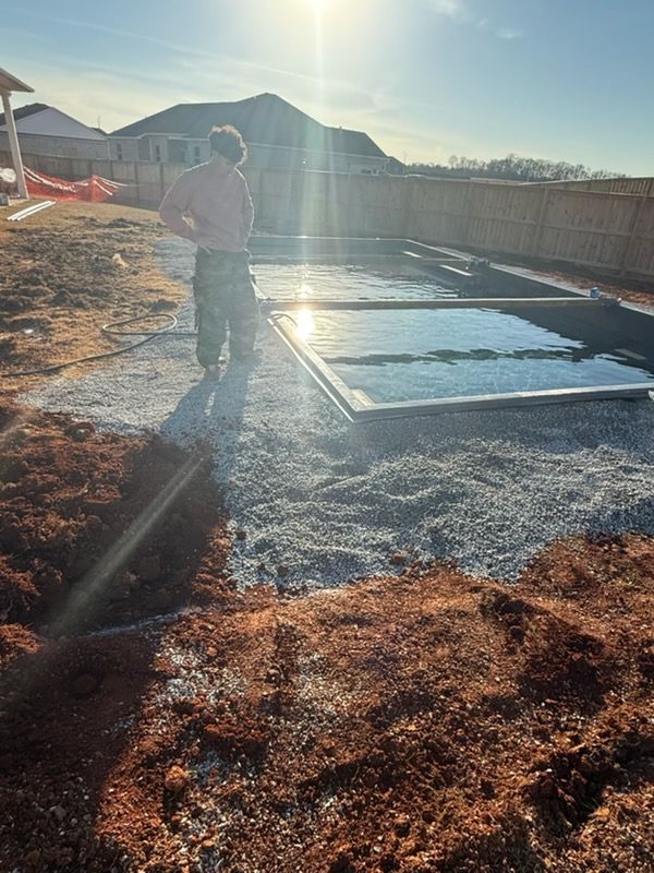Person standing on a gravel yard beside a concrete slab in bright sunlight with long shadows.