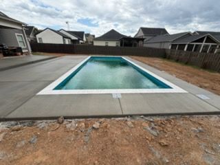 Rectangular backyard swimming pool under construction, surrounded by concrete and a dirt yard.