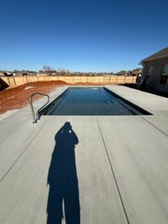 Backyard swimming pool under construction with a concrete deck, fence, and long shadow in the foreground
