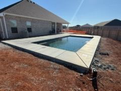 Backyard swimming pool with concrete deck beside a brick house and wooden fence under a clear sky