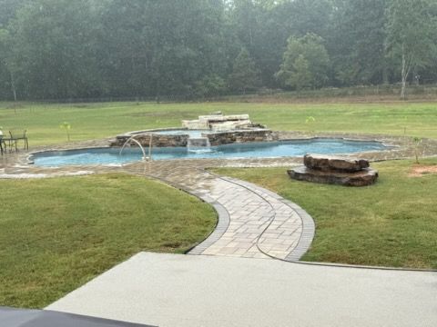 Curved stone path leading to an empty backyard pool with stone fountain, surrounded by lawn and trees.