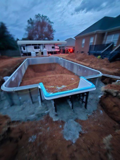 Partially built rectangular in-ground pool shell in a backyard at dusk, with houses and a tree nearby.