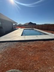 Backyard swimming pool beside a house, with concrete patio and red gravel yard under a sunny blue sky