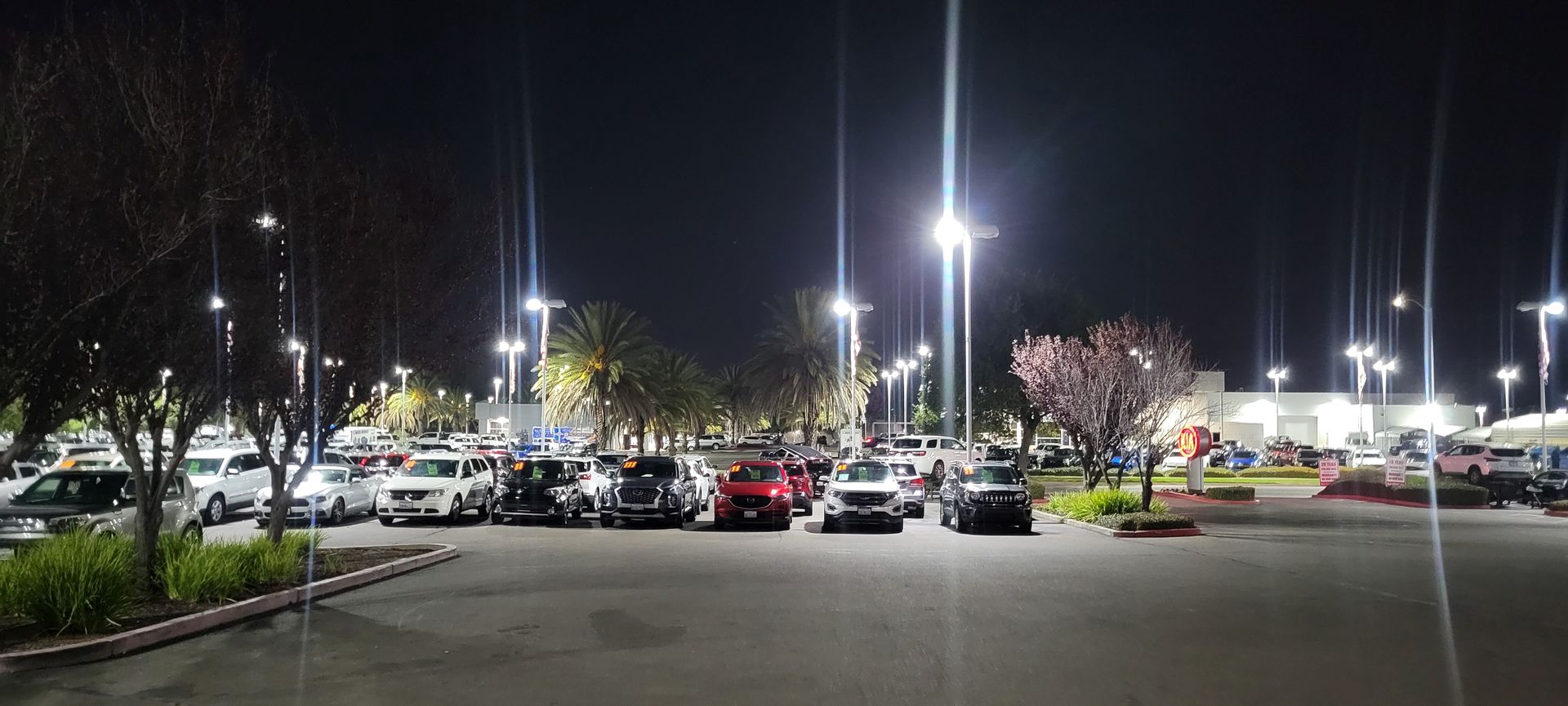 Nighttime parking lot with rows of parked cars and bright streetlights in a shopping center.