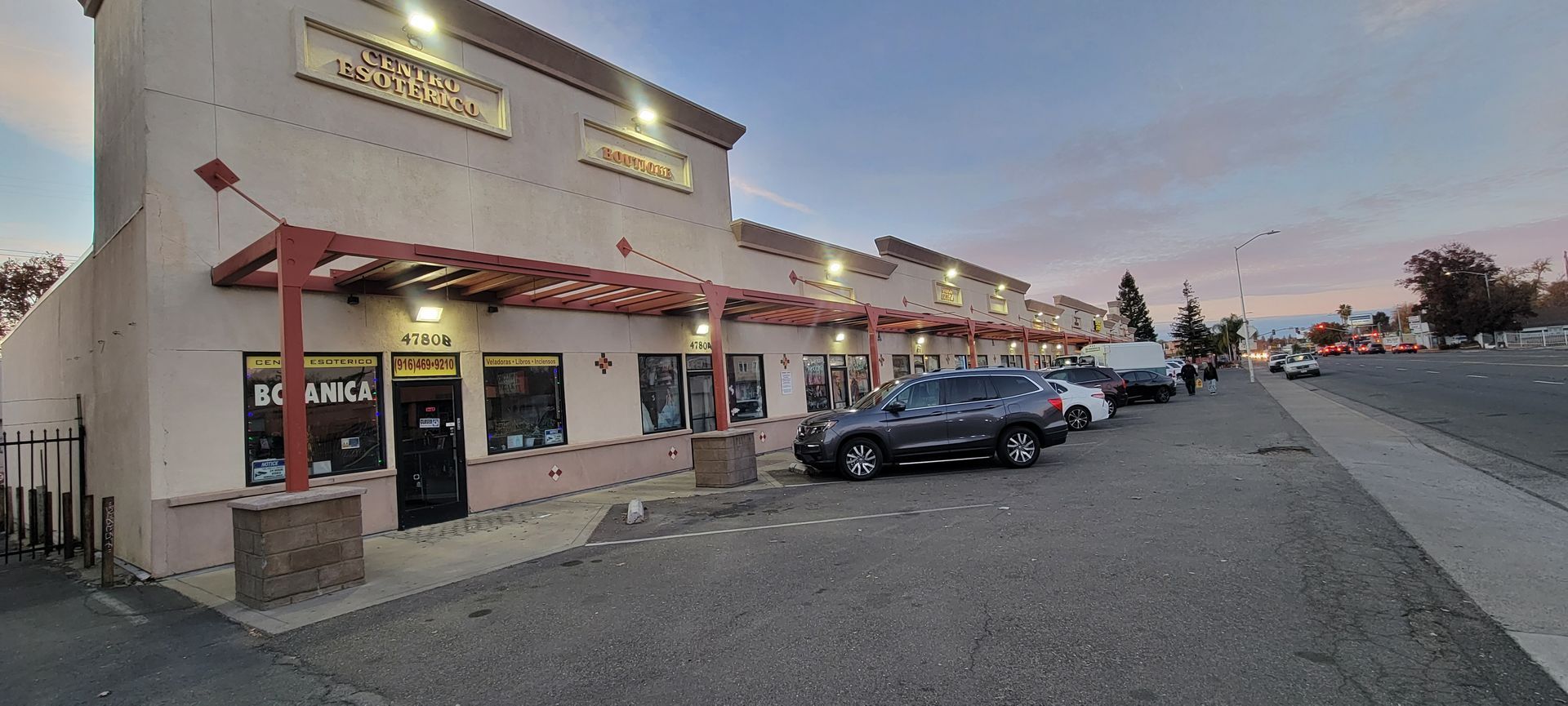 Strip mall storefront at dusk with parked cars and a red awning along the street.