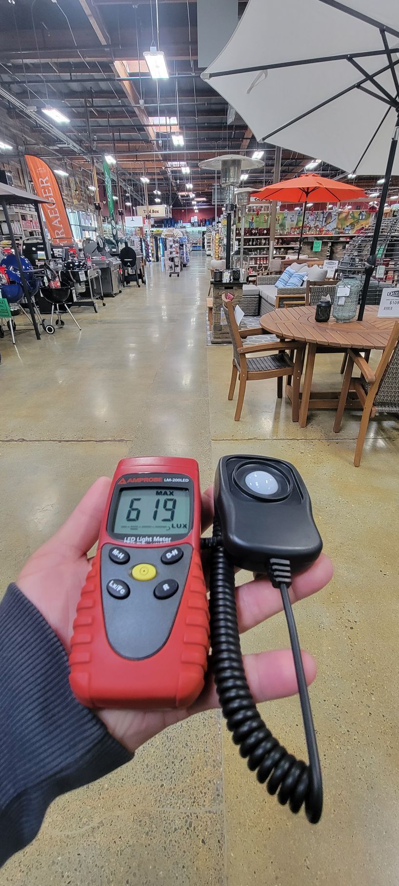 Hand holding a red digital thermometer and a black infrared gun in a warehouse store aisle.