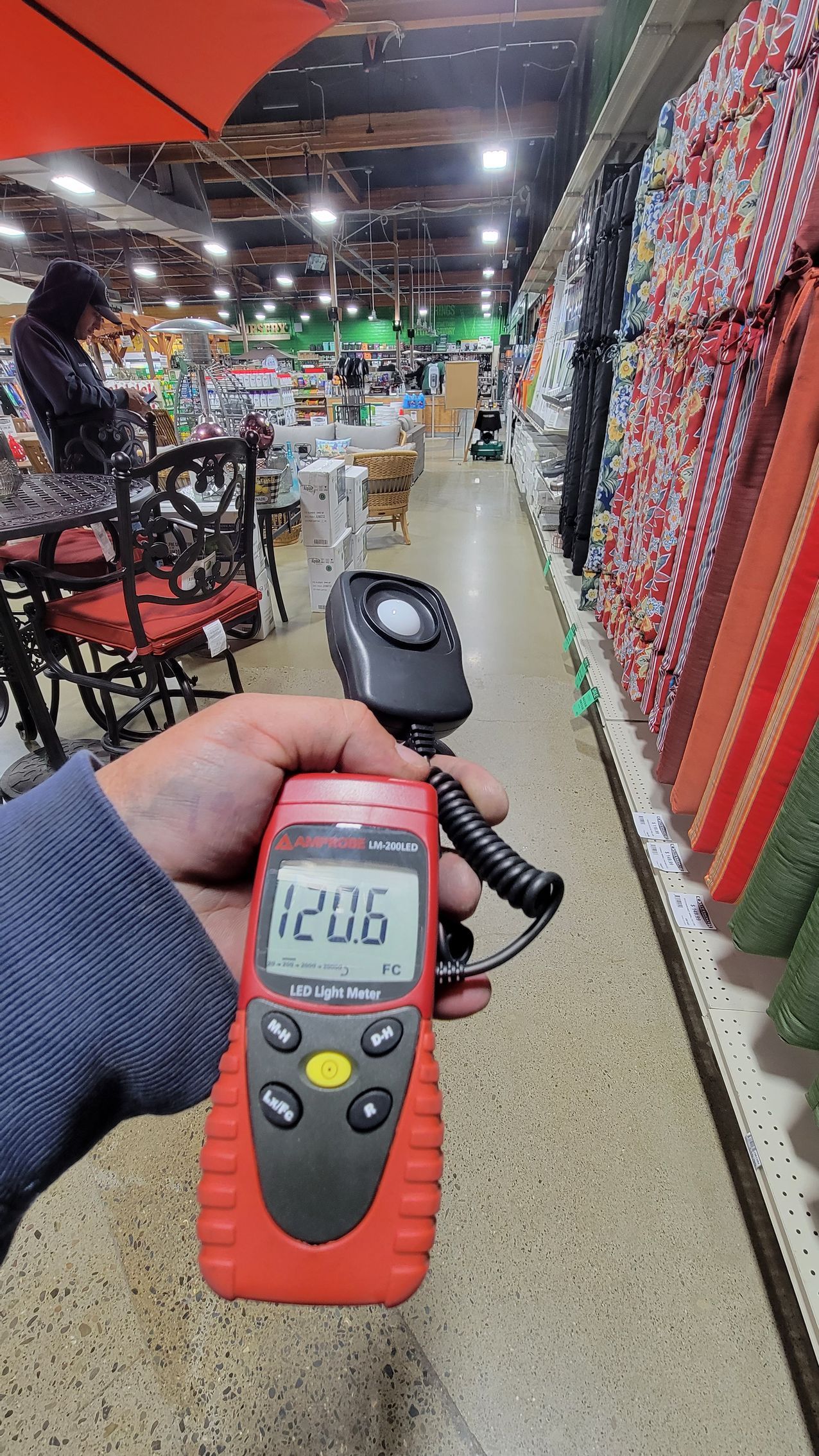 Hand holding a red handheld meter in a brightly lit shop aisle with hanging textiles on the right