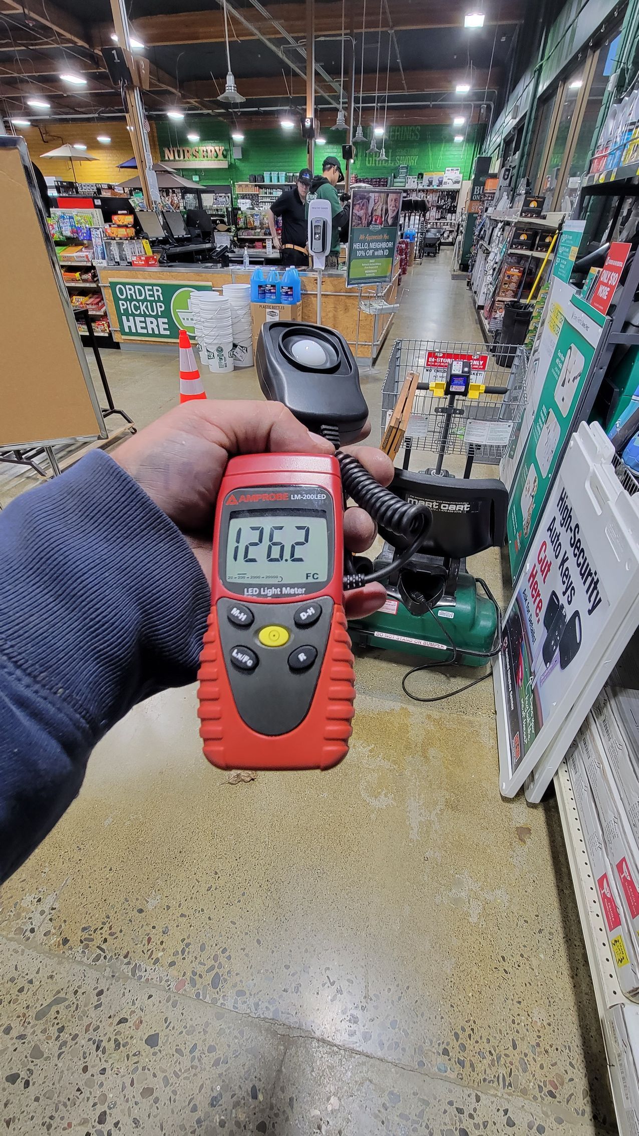 Hand holding a red digital meter in a store aisle, with shelves and tools in the background