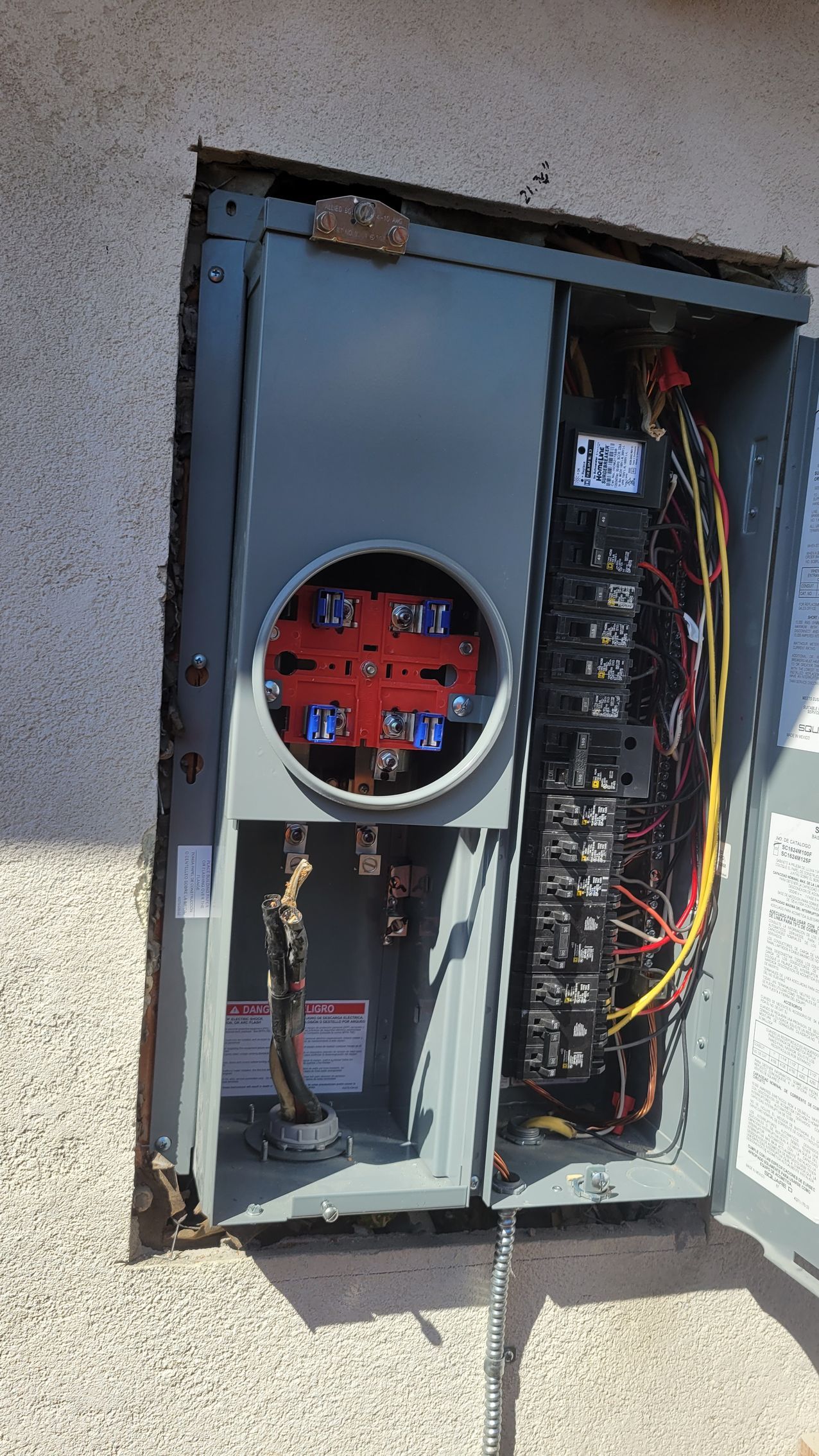Open electrical breaker panel with exposed wires and red circular meter on a stucco wall.