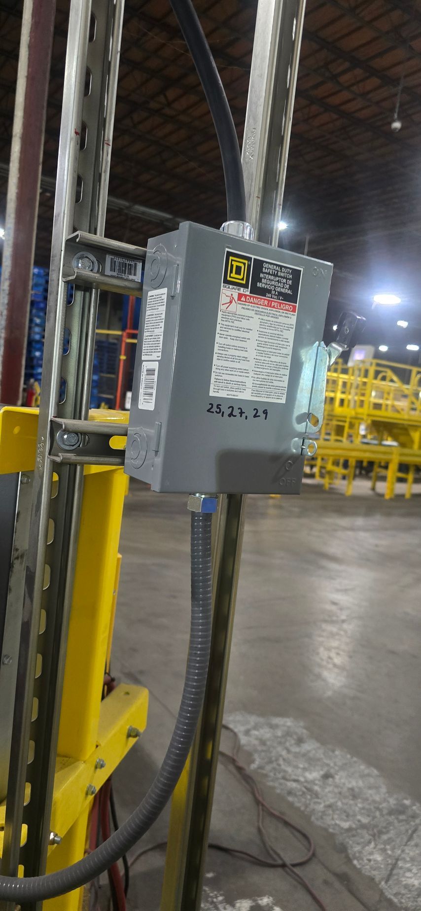 Gray electrical control box mounted on a yellow industrial post in a warehouse aisle.