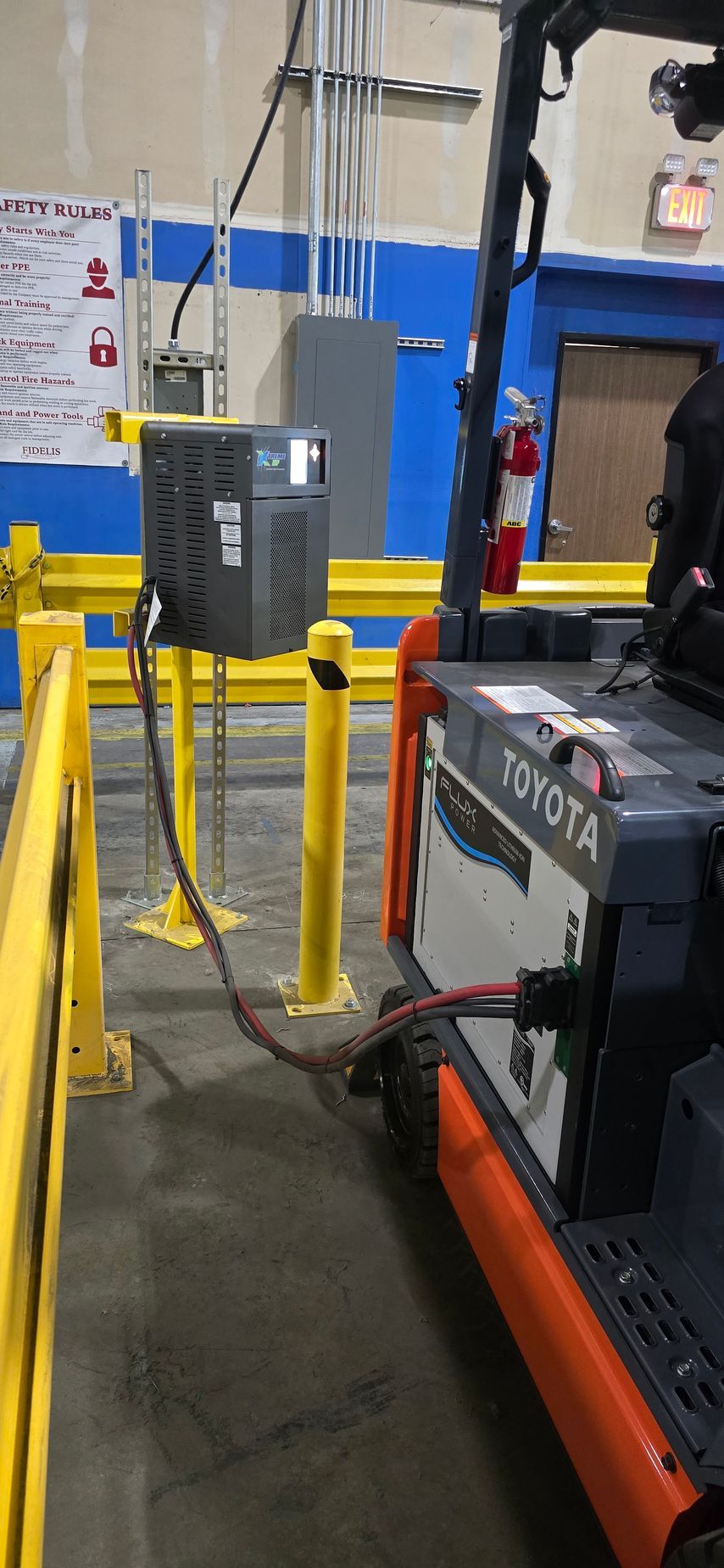 Industrial warehouse with a forklift beside a gray electrical box and yellow safety bollards.