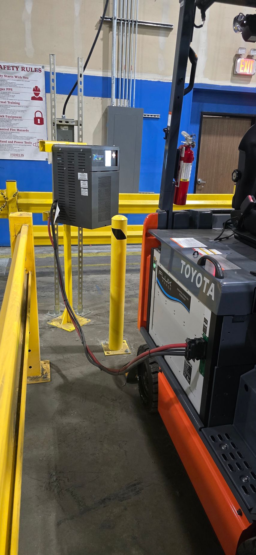 Industrial warehouse floor with yellow safety stanchions and a gray electrical box near machinery.