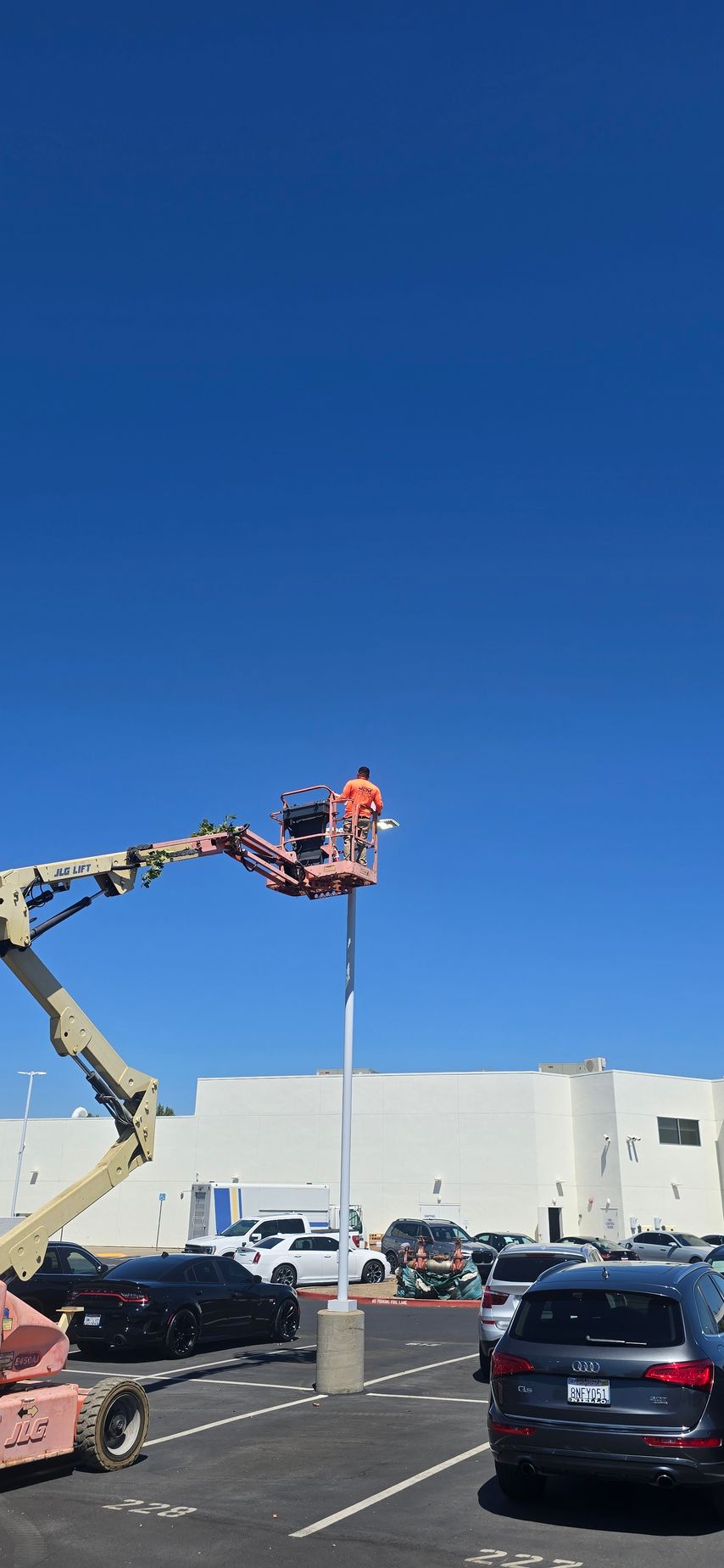 Worker on a cherry picker replacing a parking lot light pole beside a store under a clear blue sky
