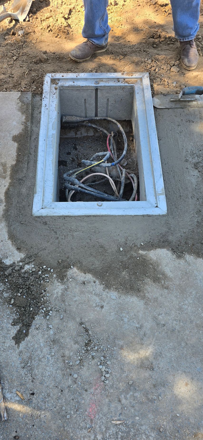 Open utility box in concrete ground with exposed cables, beside a worker’s boots