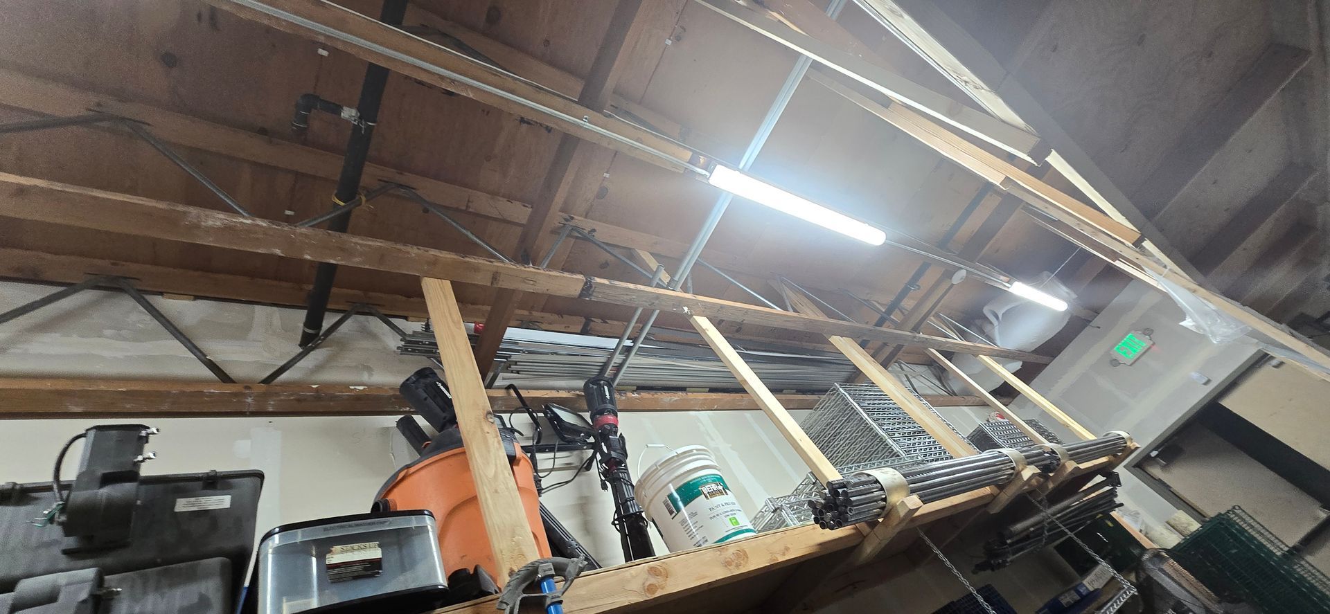 Cluttered workshop attic with shelves, tools, and exposed wooden beams under a bright fluorescent light